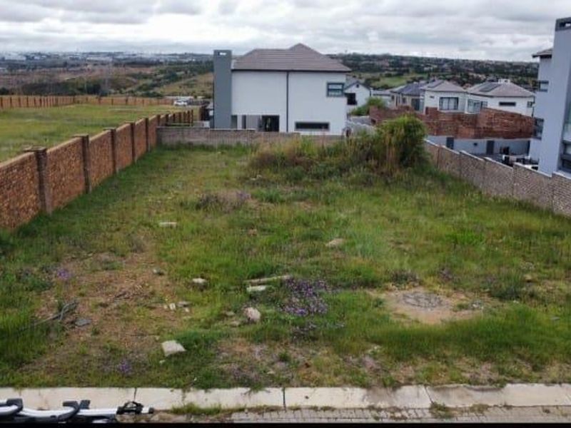A vacant residential plot featuring a brick boundary wall, grassed terrain, and established neighborhood surroundings.