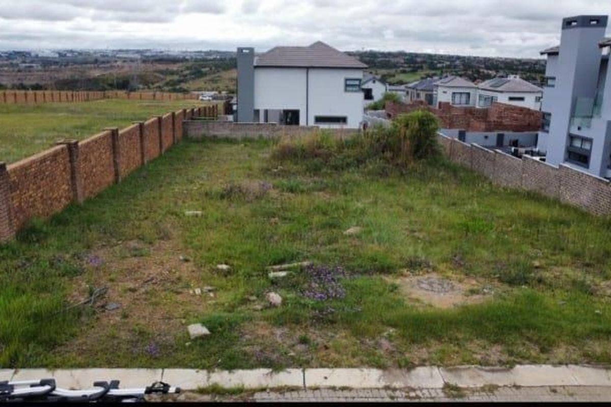 A vacant residential plot featuring a brick boundary wall, grassed terrain, and established neighborhood surroundings.