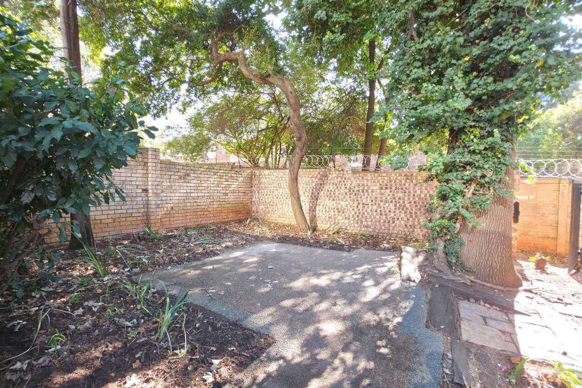 A private rear garden area featuring a concrete patio, established trees, and perimeter brick walling with security fencing.