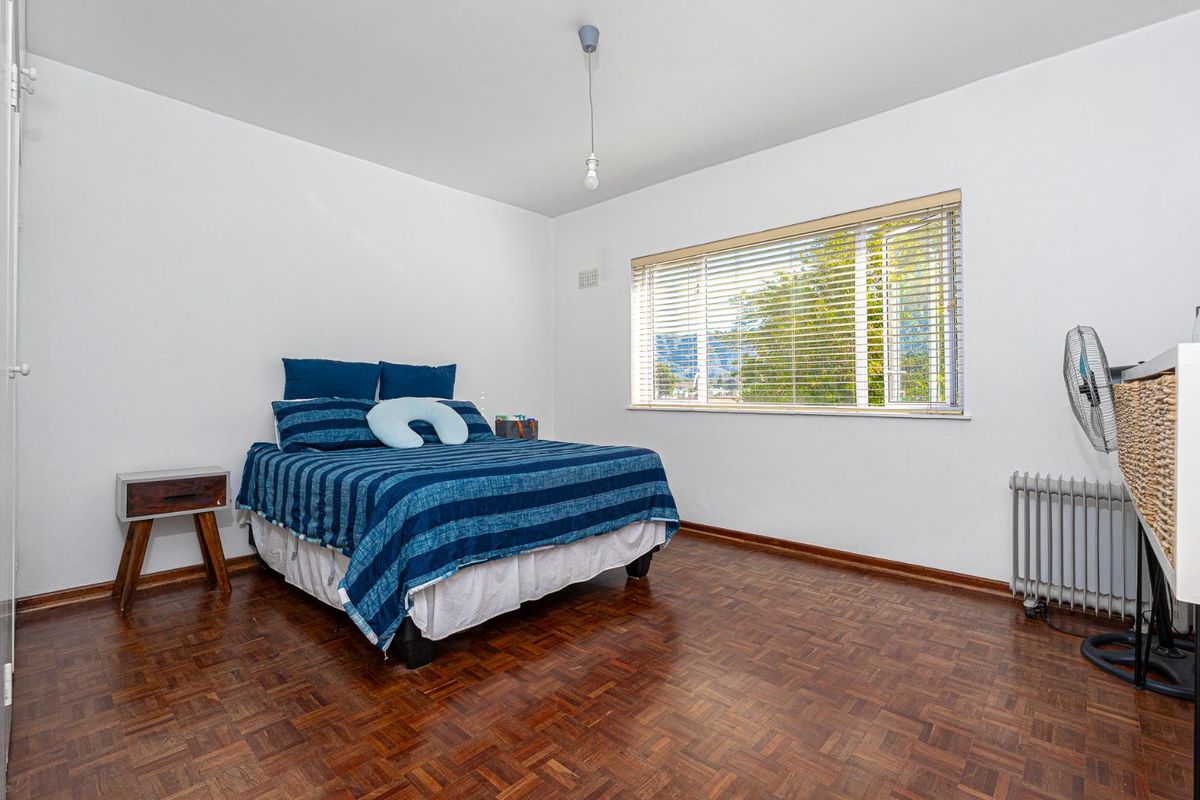 A bright bedroom featuring classic parquet flooring, a large window with fitted blinds, and a wall-mounted radiator.