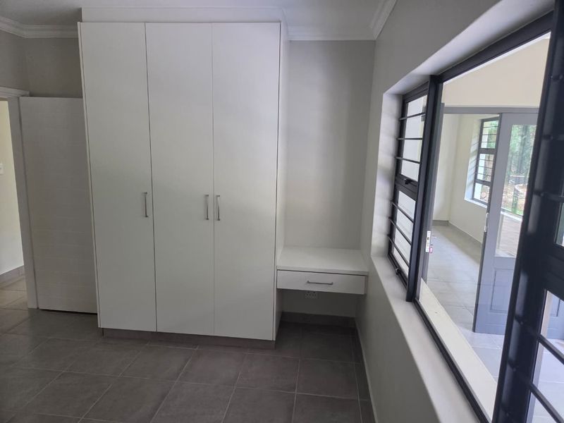 A clean, modern bedroom featuring built-in white cabinetry, a matching integrated desk, tiled floors, and large windows with security bars.