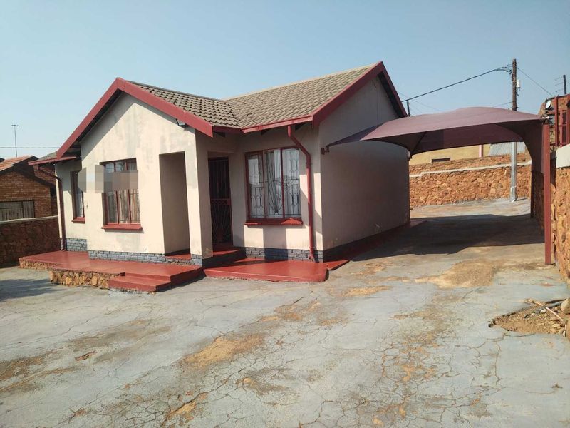 Front elevation of a single-story house featuring a tiled roof, a covered carport, and a concrete driveway with a stone boundary wall.
