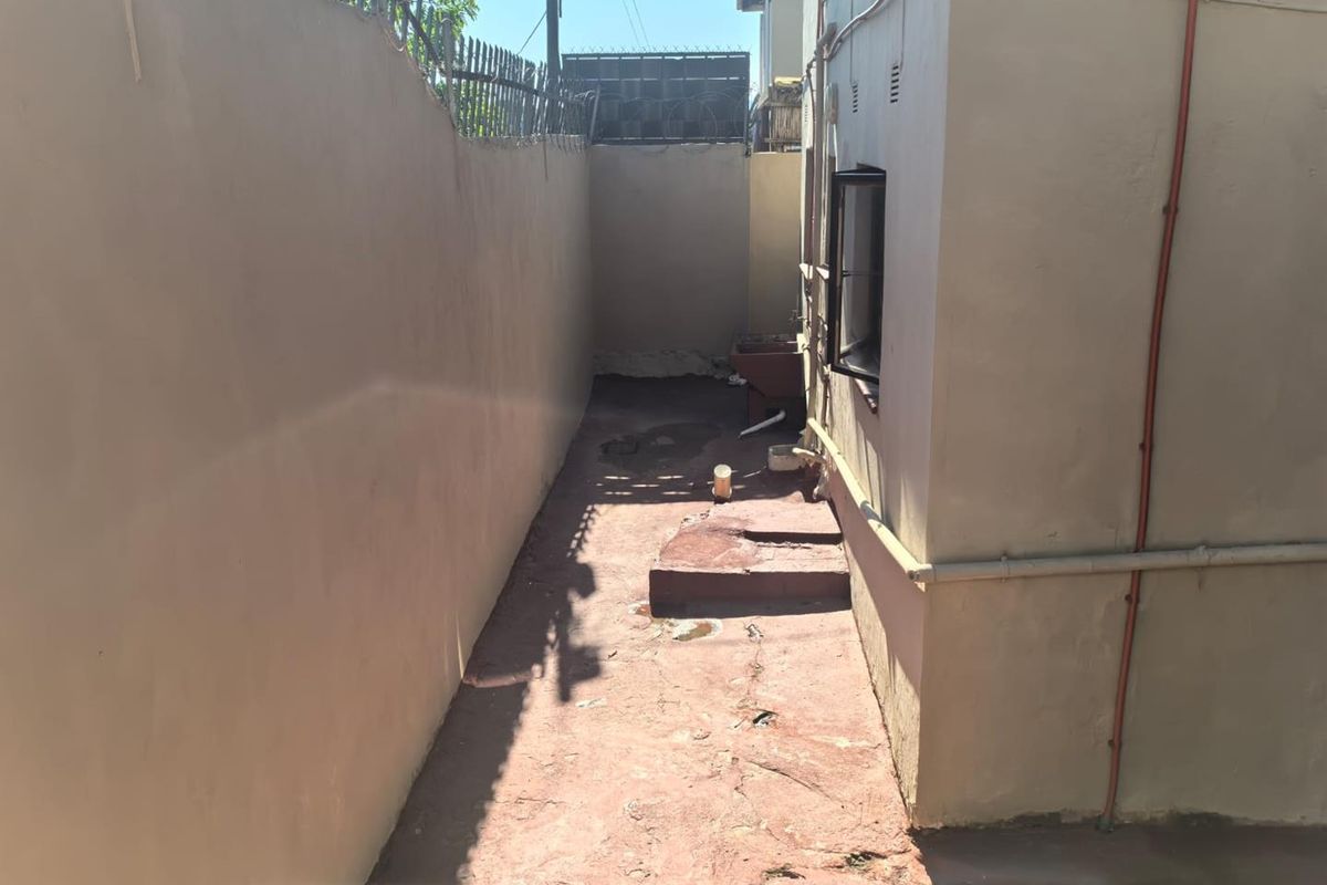 A narrow rear exterior side passage featuring concrete paving, perimeter wall with security spikes, and exposed external plumbing fixtures.