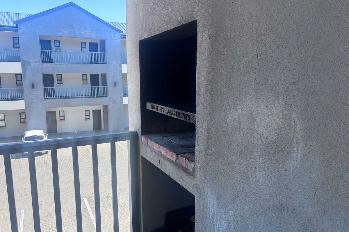 A balcony featuring a built-in braai and metal balustrade, overlooking the apartment complex courtyard and parking area.