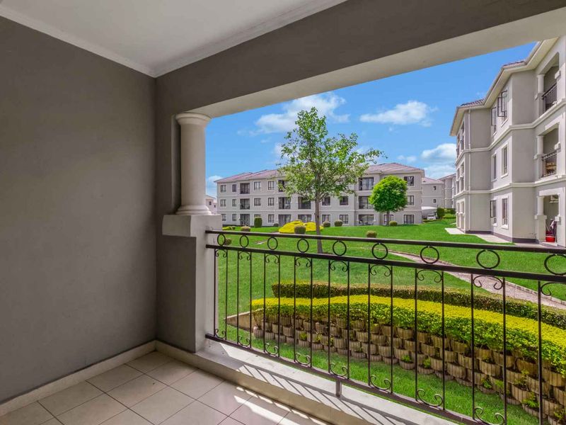 A covered balcony featuring tiled flooring and a wrought iron railing, overlooking a well-maintained communal garden and apartment complex.