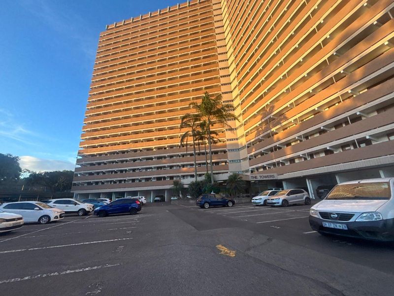 Exterior view of The Towers apartment building featuring a large asphalt parking area and multi-story residential facade with balcony access corridors.
