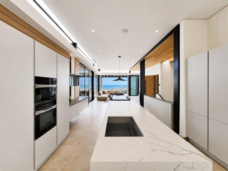 Modern open-plan kitchen and living area featuring a large stone island, high-end built-in appliances, and expansive sliding doors opening to a scenic coastal view.