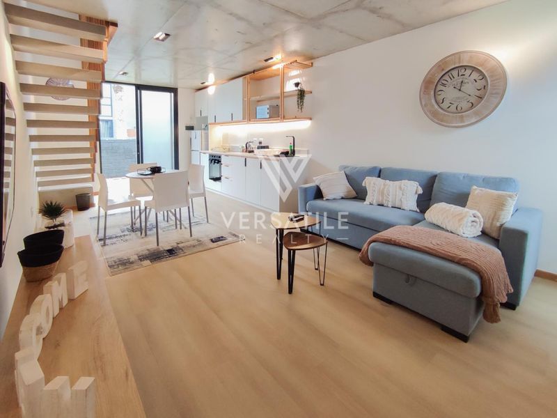 Modern open-plan living area featuring a sleek kitchen with built-in cabinetry, laminate flooring, exposed concrete ceiling, and a staircase leading to the upper level.