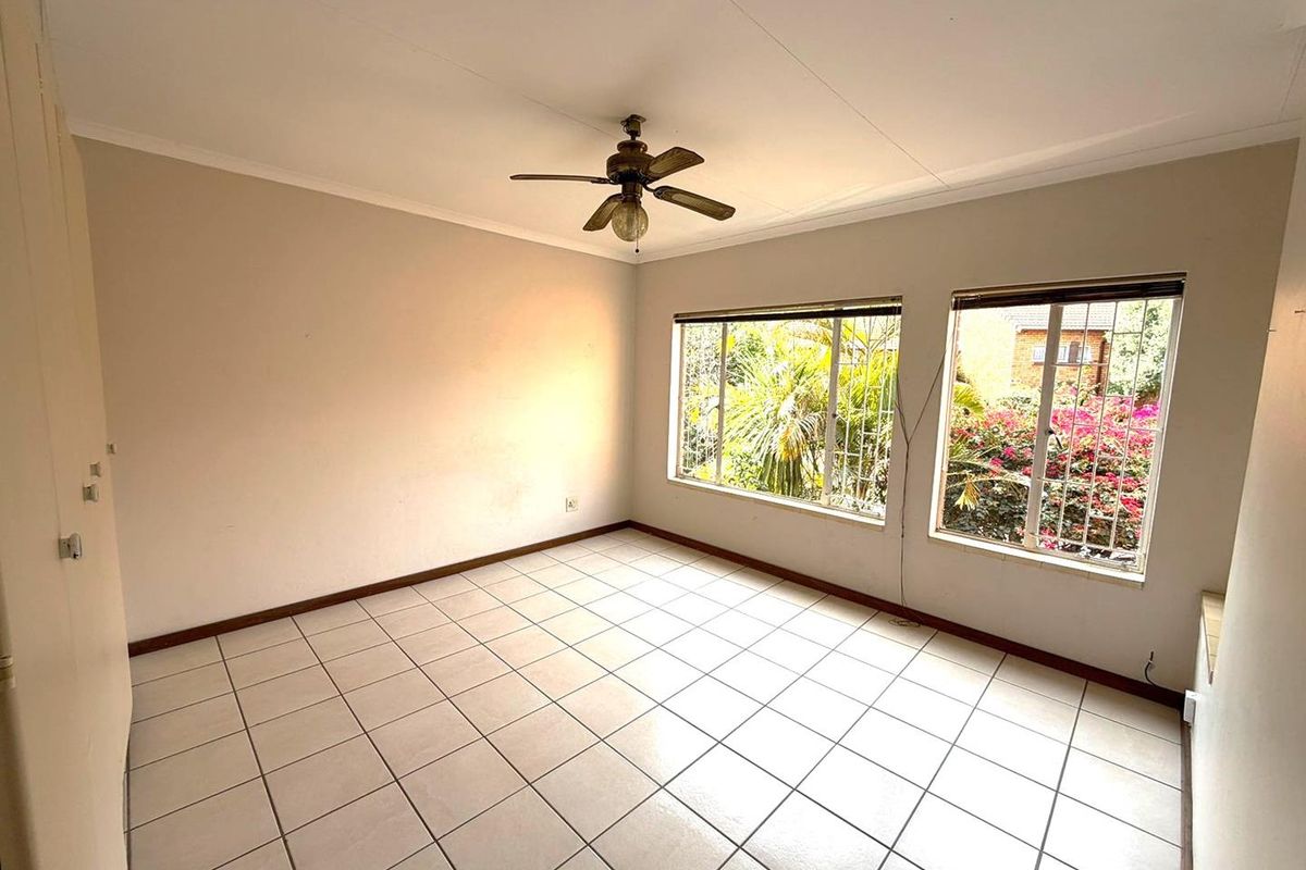 A bright, empty bedroom featuring tiled flooring, a ceiling fan, and large windows with security bars overlooking the garden.