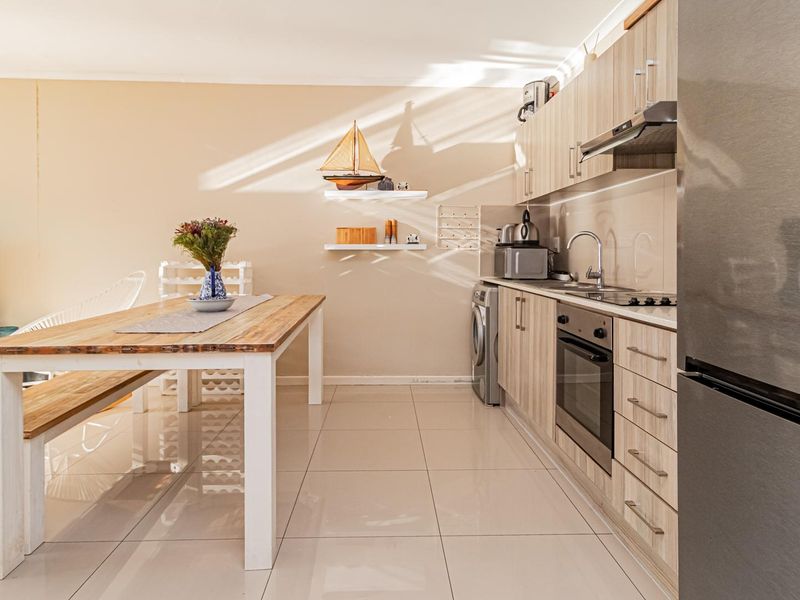 An open-plan living and kitchen area featuring modern light wood cabinetry, stainless steel appliances, and neutral tiled flooring throughout.