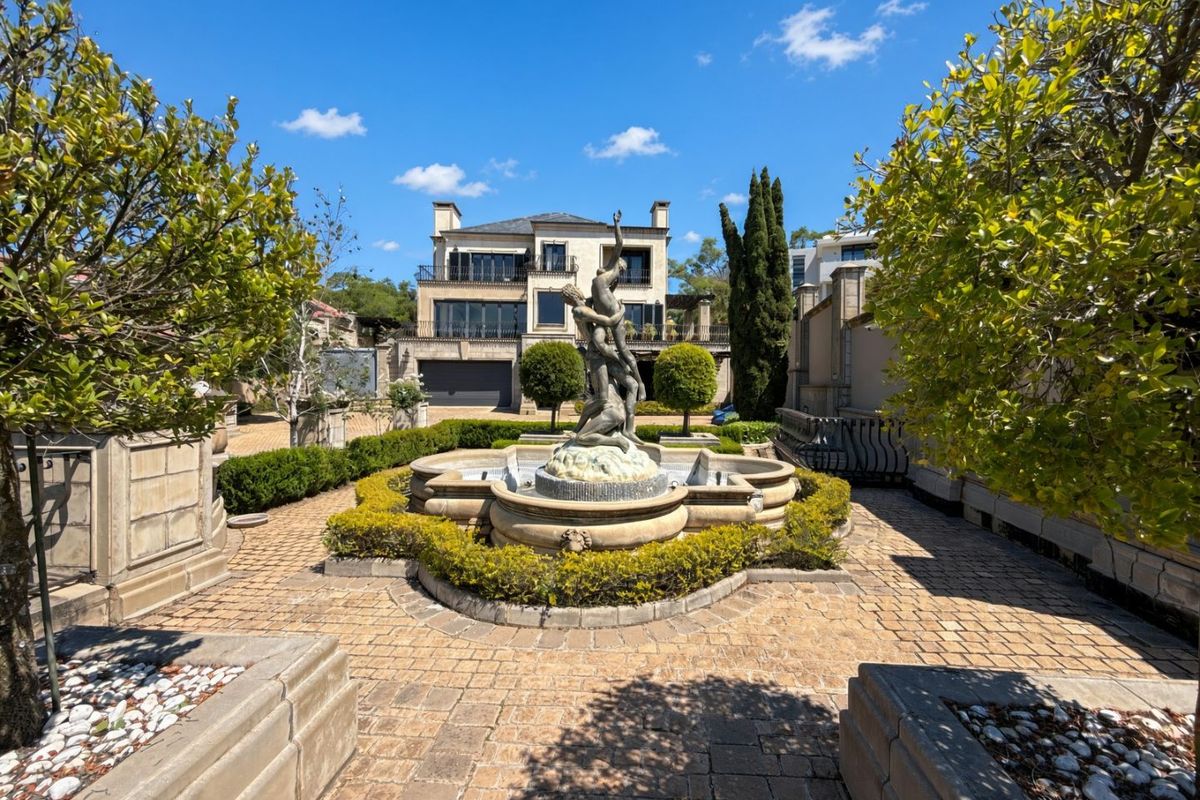 A beautifully landscaped rear courtyard featuring a central stone fountain, paved pathways, and stone balustrades, set against a multi-story residential property.