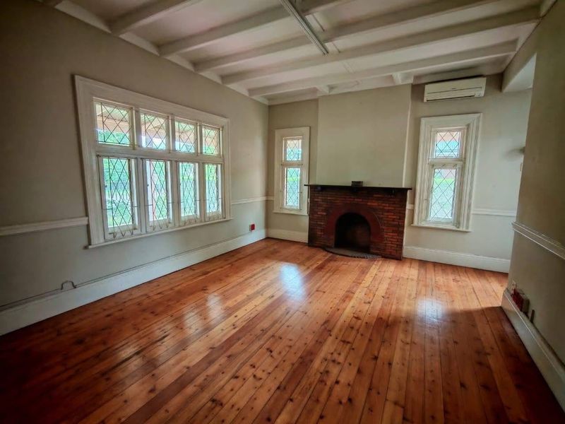 Spacious office area featuring polished timber flooring, a classic brick fireplace, exposed beam ceiling, and large leaded glass windows providing ample natural light.