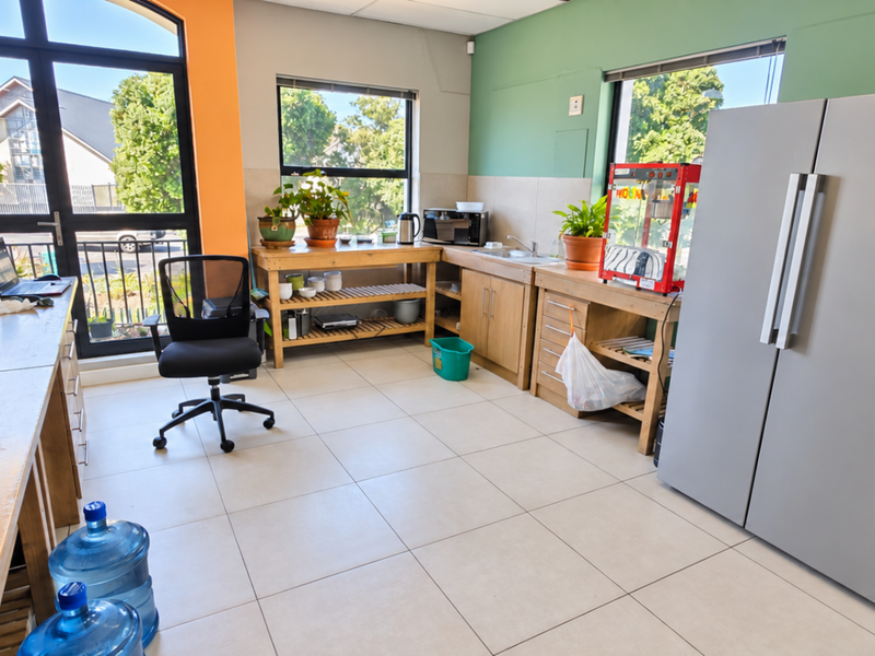 A functional office kitchen area featuring tiled flooring, built-in wooden cabinetry, a stainless steel sink, and large windows providing ample natural light.