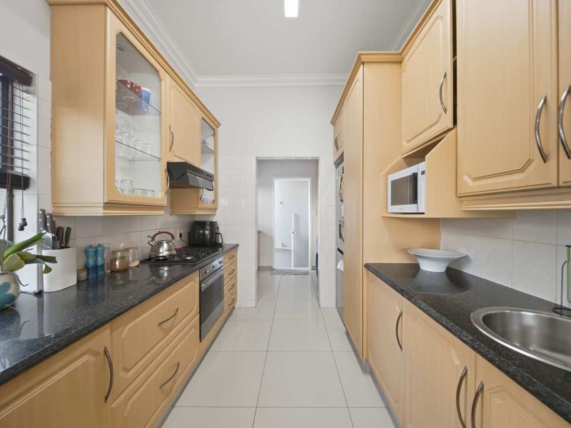 A functional galley-style kitchen featuring light wood cabinetry, dark granite countertops, built-in appliances, and tiled flooring, leading to a separate utility area.