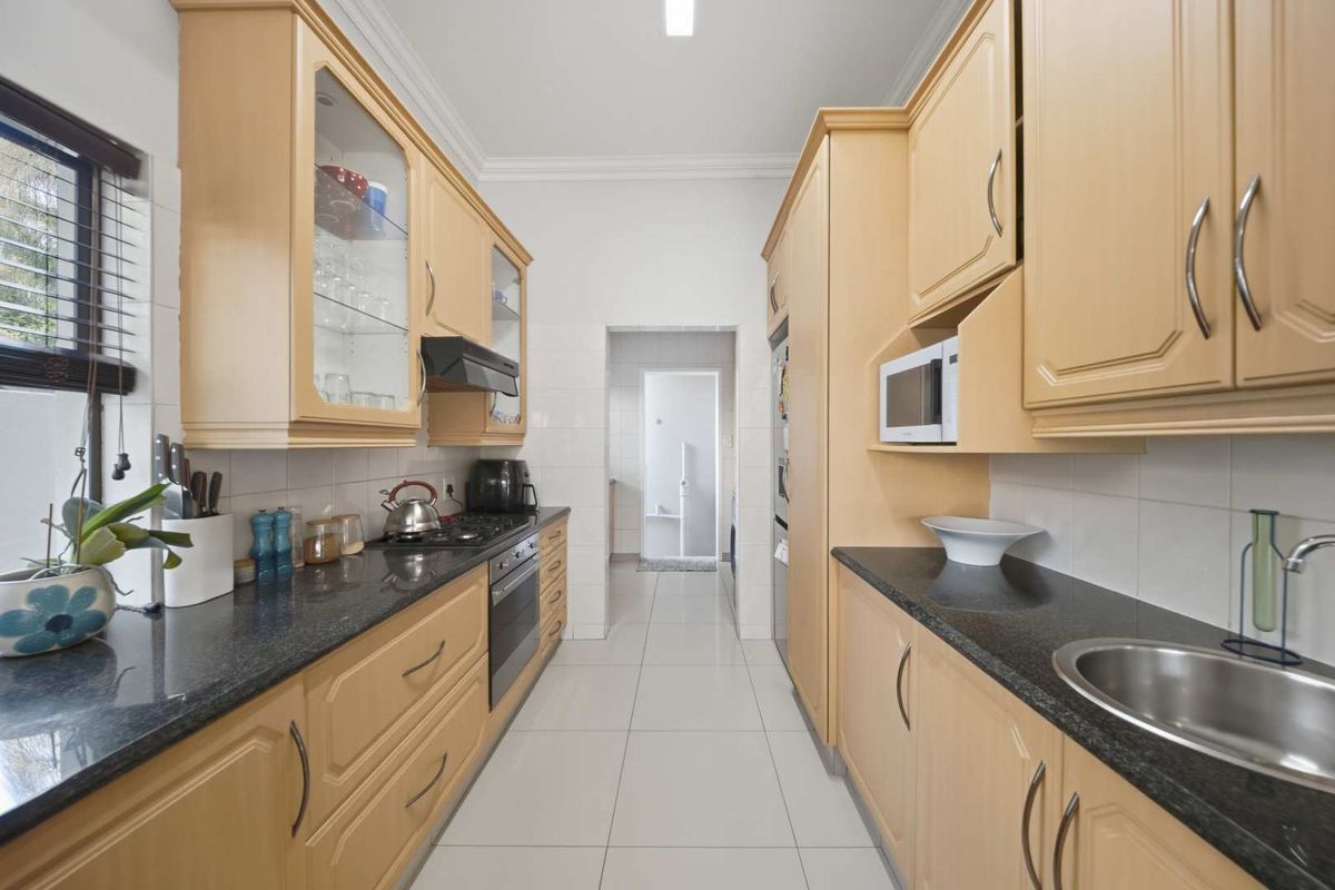 A functional galley-style kitchen featuring light wood cabinetry, dark granite countertops, built-in appliances, and tiled flooring, leading to a separate utility area.