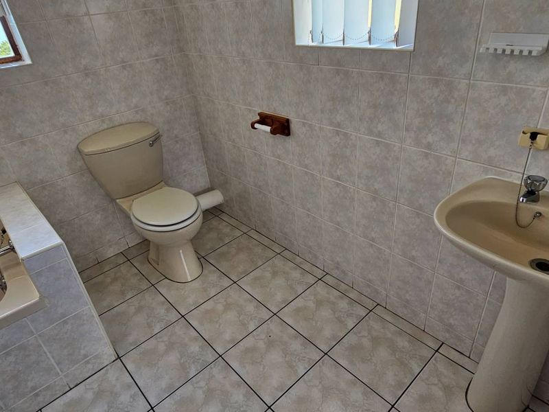 A dated bathroom featuring a built-in bathtub, pedestal basin, and toilet, finished with neutral wall and floor tiles.