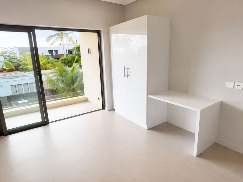 A bright, modern bedroom featuring neutral tiled flooring, a built-in wardrobe with an integrated desk, and large sliding glass doors opening to a balcony.