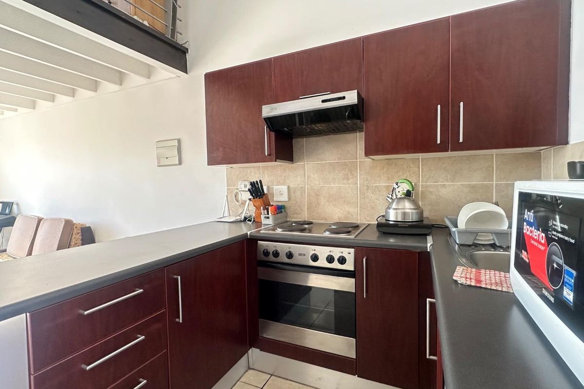 Compact kitchen featuring dark wood cabinetry, stainless steel oven, electric hob, extractor fan, and tiled backsplash, situated beneath a mezzanine level.