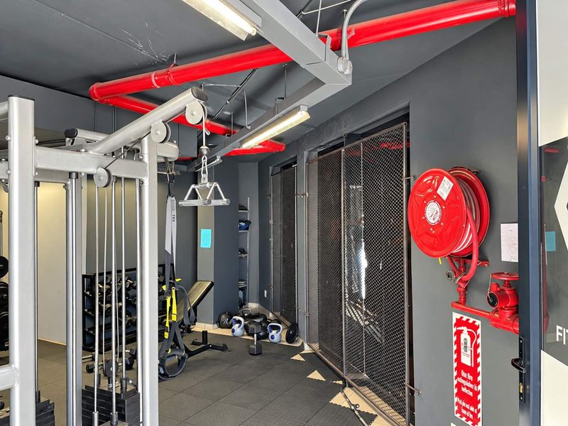 A functional gym space featuring industrial-style exposed red fire piping, rubber flooring, metal mesh partitions, and integrated fire safety equipment.