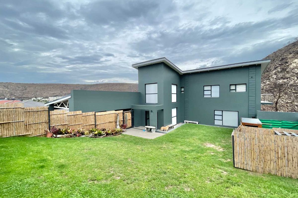 Rear view of a modern two-story house featuring dark green exterior walls, large windows, and a paved patio area leading to a grass lawn.