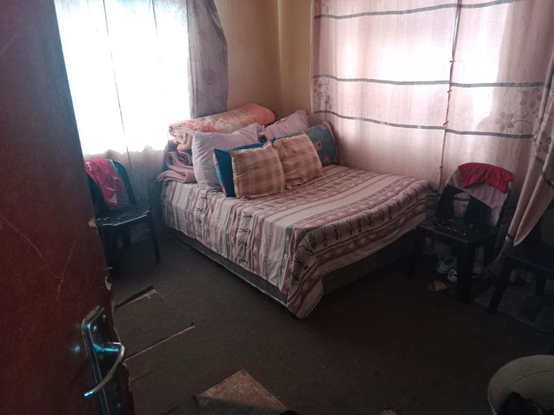 A compact bedroom featuring natural light through windows with curtain rails and worn flooring, requiring cosmetic updates to improve the overall living space.