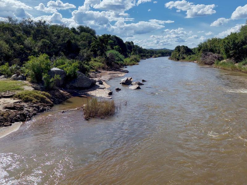 A scenic view of a flowing river surrounded by natural vegetation and rock formations under a bright blue sky.