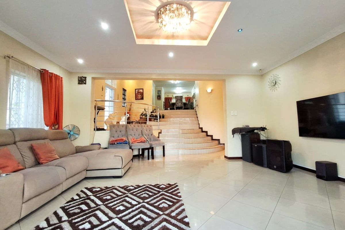 Spacious living room featuring tiled flooring, a recessed ceiling with a central chandelier, and an integrated staircase with a stainless steel balustrade.
