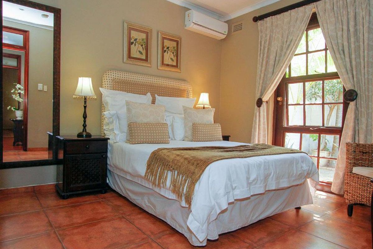 A cozy bedroom featuring terracotta floor tiles, a wall-mounted air conditioning unit, and a large wooden-framed window with garden views.