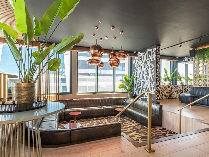 A modern, stylish lounge area featuring a sunken seating section, large windows, contemporary pendant lighting, and unique wall art in a sophisticated apartment building.