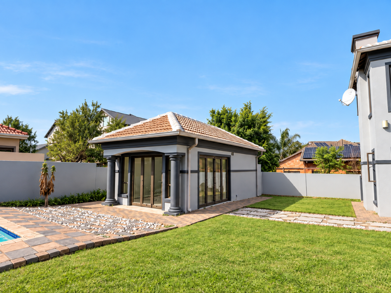 Rear exterior view featuring a swimming pool, paved deck, landscaped lawn, and a detached structure with sliding glass doors and decorative columns.
