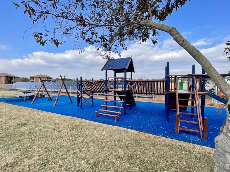 Outdoor communal playground area featuring wooden climbing structures, swings, and blue safety flooring, situated near a solar panel installation.
