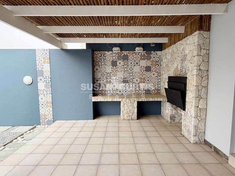 A well-maintained covered patio featuring a built-in braai, decorative tiled accents, stone cladding, and a rustic thatched roof structure.