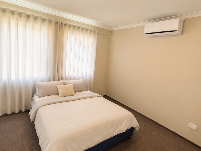 A well-maintained bedroom featuring neutral wall tones, carpeted flooring, a wall-mounted air conditioning unit, and built-in wardrobe storage.