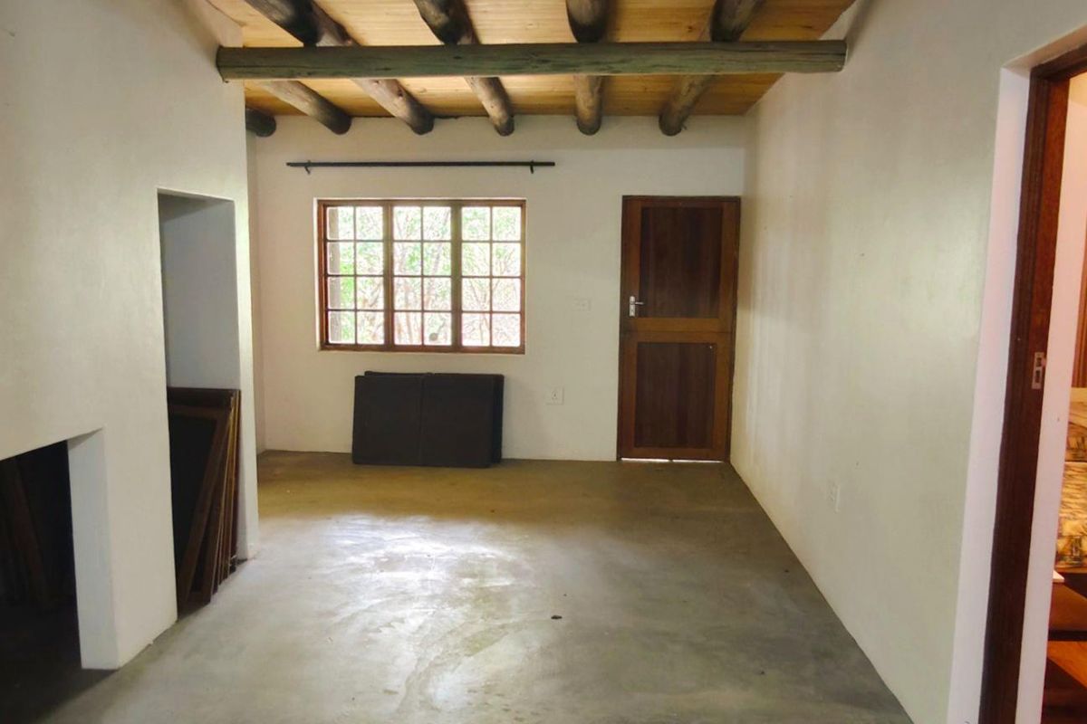 An interior view of a room featuring exposed timber ceiling beams, a wooden door, and a window, with concrete flooring throughout.