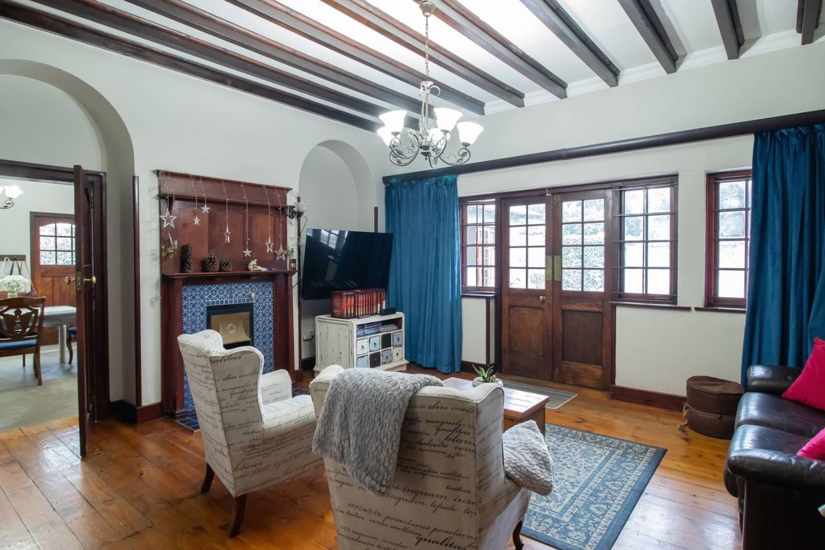A cozy living room featuring exposed timber ceiling beams, polished timber flooring, a tiled fireplace with a wooden mantel, and classic timber-framed french doors.