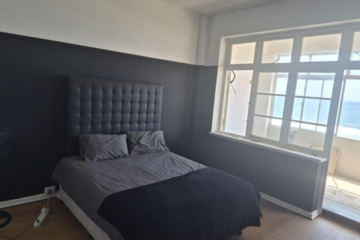 A bedroom featuring timber flooring and a large window with direct access to a balcony, offering natural light and ocean views.