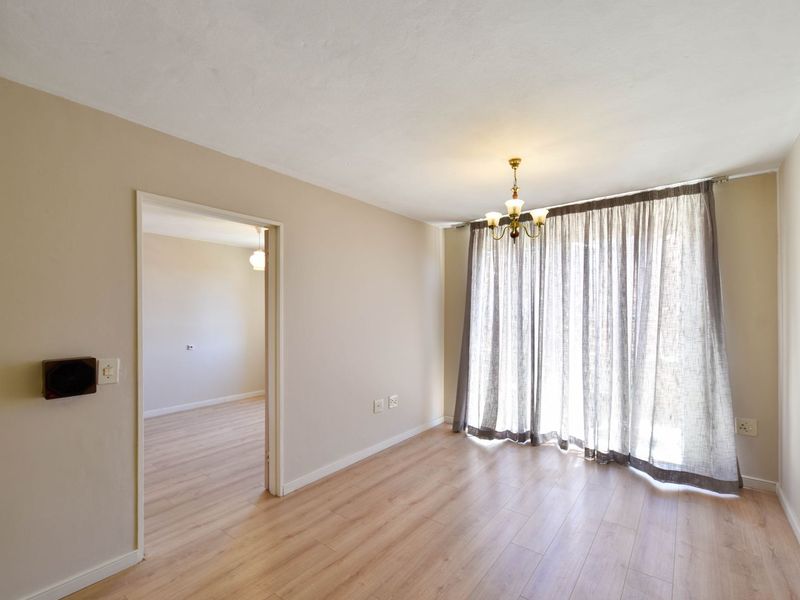 A bright, empty living room featuring light-toned laminate flooring, neutral walls, and a ceiling-mounted light fixture, with a doorway leading to an adjacent room.