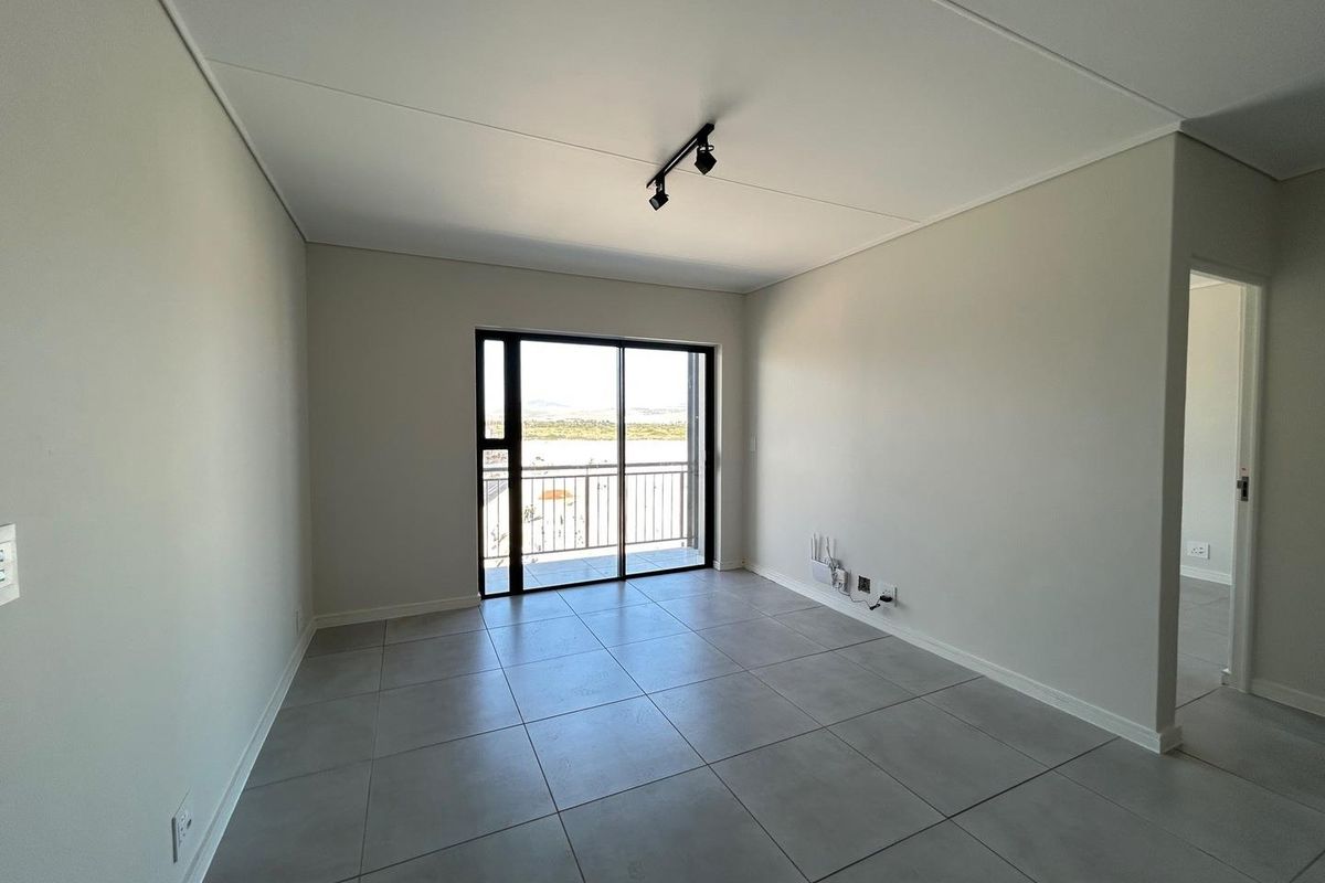 A bright, empty living area featuring large format grey floor tiles, modern track lighting, and sliding glass doors leading to a balcony.