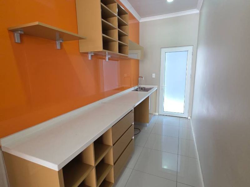 A clean, modern galley-style kitchen featuring vibrant orange glass splashbacks, ample built-in cabinetry, white countertops, and polished floor tiles.