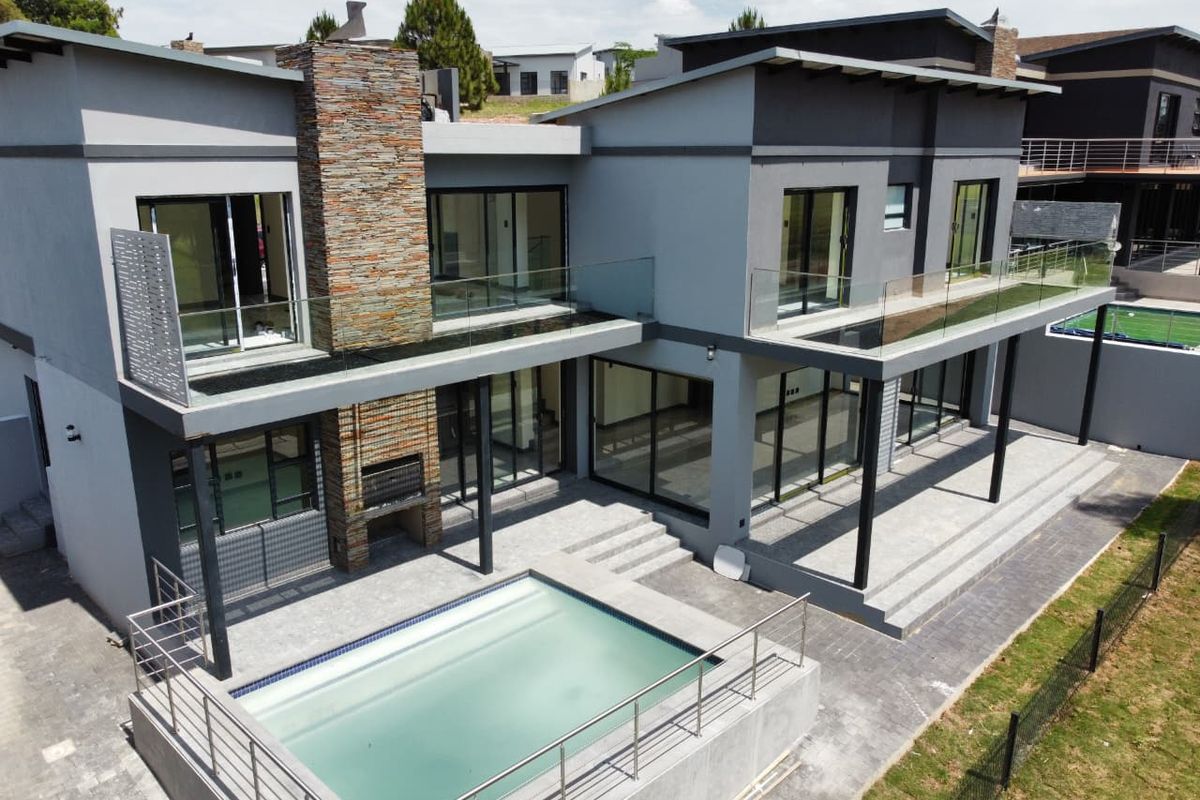 Aerial view of a modern double-story cluster home featuring a swimming pool, stone-clad chimney, built-in braai, and expansive upper-level balconies with glass balustrades.