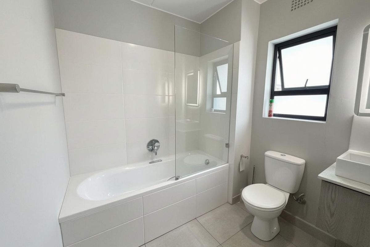A modern, clean bathroom featuring a built-in bathtub with a glass shower screen, a white toilet, and a vanity unit with a basin.