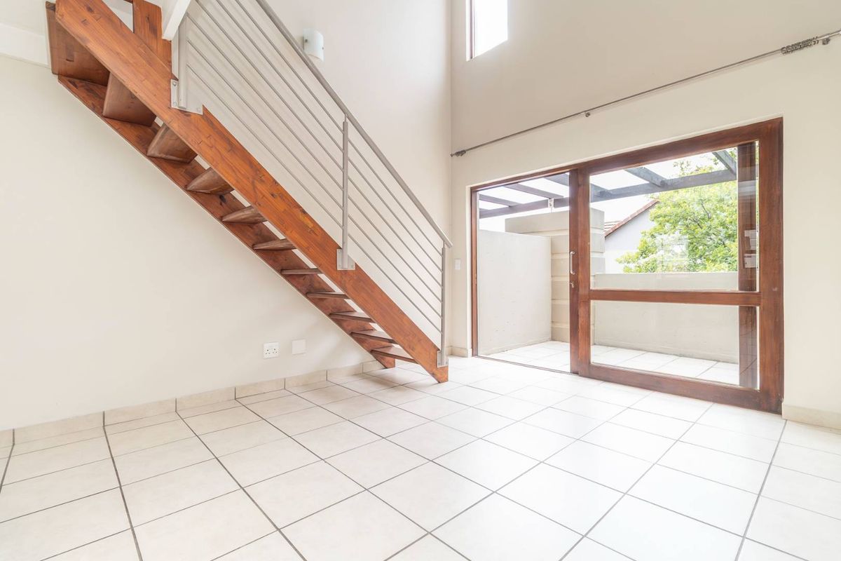 Bright, double-volume living area featuring tiled floors, a wooden staircase with metal balustrade, and direct access to an outdoor patio through sliding glass doors.