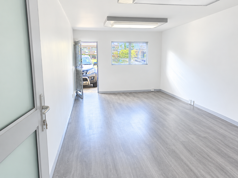 A bright, empty office space featuring modern laminate flooring, clean white walls, and a large window with venetian blinds, providing a versatile workspace.