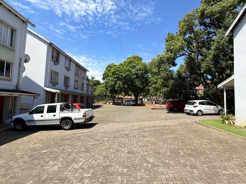 Exterior view of an apartment complex featuring a paved parking area and mature landscaping between the residential buildings.