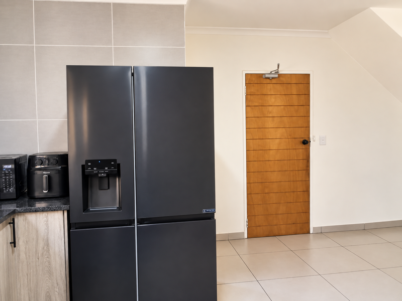 A modern kitchen area featuring tiled flooring, a tiled splashback, granite countertops, and a solid wooden door with a functional door closer.