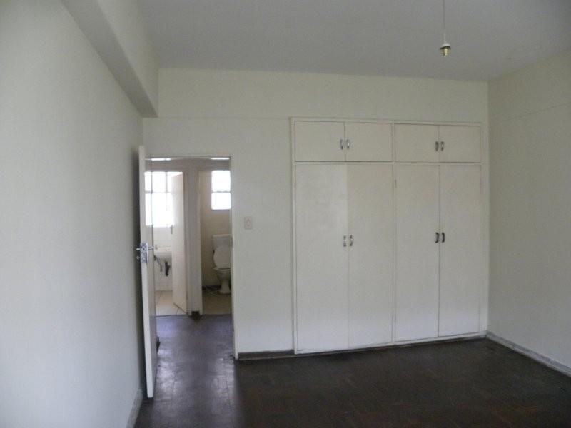 An empty bedroom featuring built-in cupboards and original parquet flooring, with an adjacent view into a bathroom area.