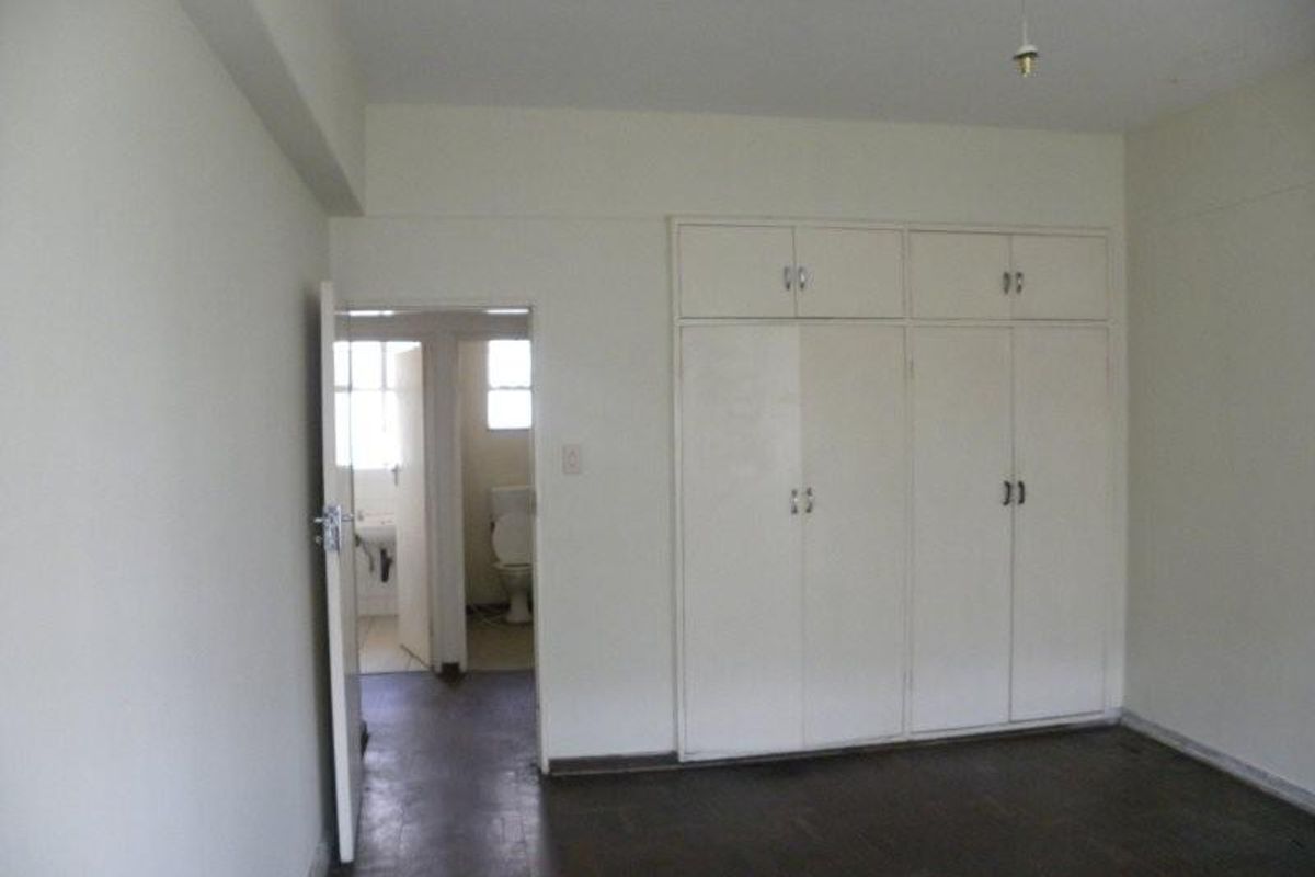 An empty bedroom featuring built-in cupboards and original parquet flooring, with an adjacent view into a bathroom area.