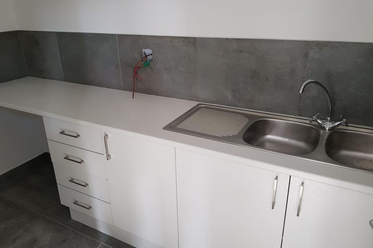 A functional kitchenette area featuring white cabinetry, a stainless steel double sink, and grey tiled splashback with exposed electrical wiring.