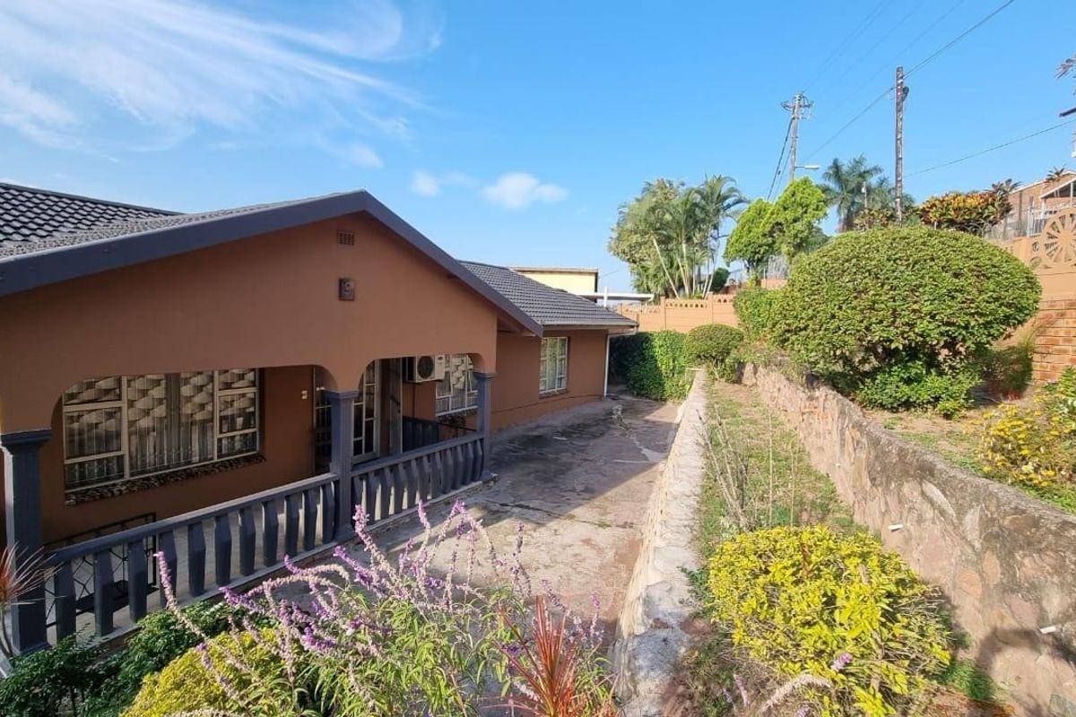 Rear exterior view of the house featuring a covered patio, concrete paving, and a stone retaining wall set against a landscaped garden.