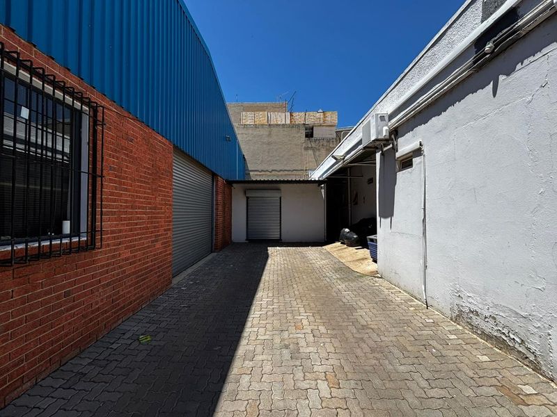 A paved rear service alleyway between two industrial buildings, featuring roller shutter access doors and security-barred windows.